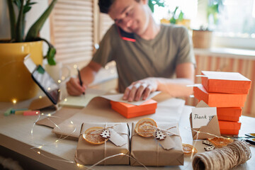Young millennial worker packing Christmas gift boxes for clients, family small business. New Year...