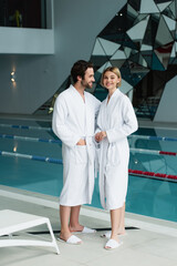 Smiling couple in bathrobes standing near swimming pool in spa center