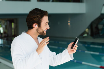 Side view of smiling man in earphone having video call on smartphone in spa center