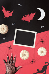 Abstract Halloween composition in red, black and white. Copy-space on blackboard. Paper silhouettes of Moon, bats, ghosts, eyes. Hand supports pyramid of decorative pumpkins. Balance concept mockup.