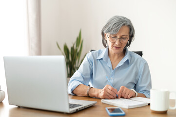 Serious mature senior female office worker business lady listening to an online webinar on laptop, taking notes in notebook, working on freelance project at home, remote work, learning at home concept