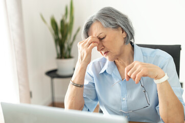 Tired and overworked stressed mature woman working on laptop at home indoor, female teacher feeling exhausted after lecturing students online, touching eyes. Smart working senior people and technology