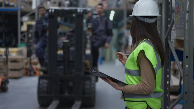 Concentrated female freight agent in hard hat standing in warehouse filling in bill of lading as men passing on forklift. Portrait of slim beautiful Caucasian woman working in industrial storehouse