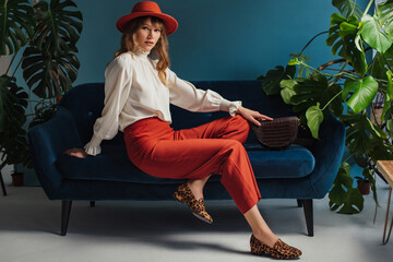 Autumn fashion conception: elegant lady wearing orange hat, vintage style blouse, trendy culottes, leopard print loafer shoes, holding stylish brown bag, posing on sofa, in blue interior