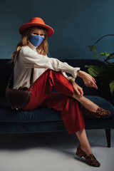 Autumn fashion portrait of elegant woman wearing trendy autumn outfit with face mask, orange hat, culottes, vintage style blouse, leopard print loafers, holding brown bag, sitting, posing on blue sofa
