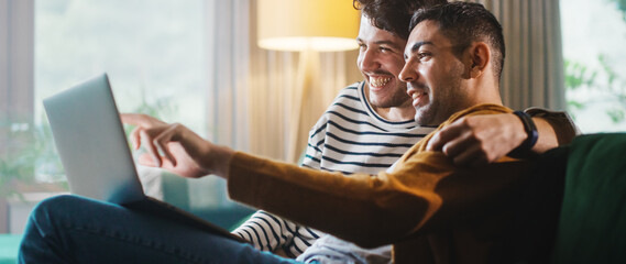 Portrait of Gentle Gay Couple Using Laptop Computer, while Sitting on a Couch in Cozy Stylish...