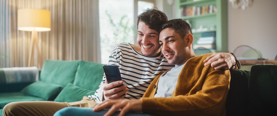 Portrait of Gentle Gay Couple Using Smartphone, while Sitting on a Couch in Cozy Stylish Apartment....