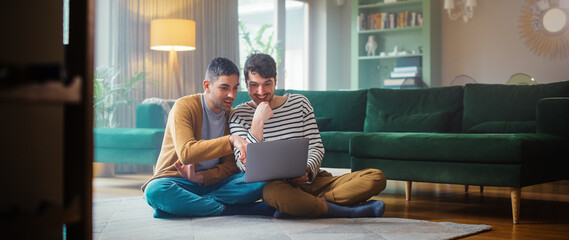 Handsome Gay Couple Using Laptop Computer, while Sitting on a Living Room Floor in Cozy Stylish...