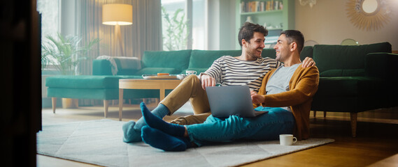Handsome Gay Couple Using Laptop Computer, while Sitting on a Living Room Floor in Cozy Stylish...