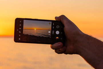 Hand holding a mobile phone and making photo of sunset over the sea. Man hand holding phone during...