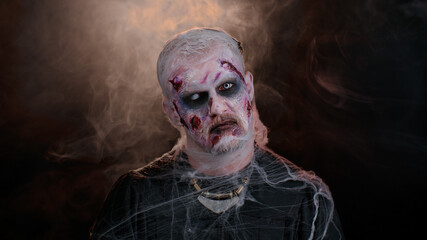 Frightening man with Halloween zombie bloody wounded makeup, trying to scare, face expressions. Convulsions. Sinister undead guy isolated on studio black background. Voodoo rituals. Fashion body art