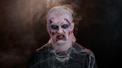 Sinister man with horrible scary Halloween zombie makeup in convulsions making faces, looking ominous, trying to scare. Dead guy with wounded bloody scars face isolated against black wall background