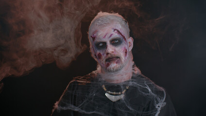 Sinister man with horrible scary Halloween zombie makeup in convulsions making faces, looking ominous, trying to scare. Dead guy with wounded bloody scars face isolated against black wall background