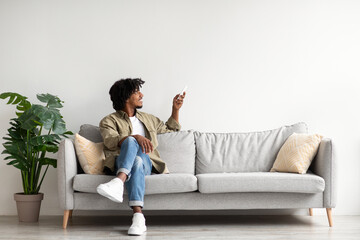 Happy black guy relaxing on couch, opening air conditioner with remote controller