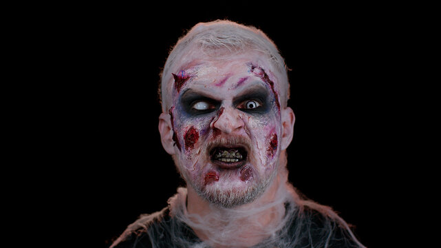 Zombie man with makeup with fake wounds scars and white contact lenses looking at camera and clicks his teeth, trying to scare in dark room. Sinister dead guy. Halloween, filming, staging concept