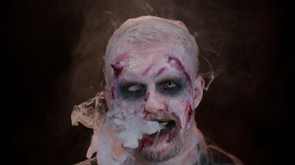 Sinister man with horrible scary Halloween zombie makeup in costume looking ominous at camera blows...