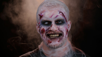 Creepy man with bloody scars face, Halloween stylish zombie make-up. Scary wounded undead guy making faces, looks at camera and smiles terribly. Voodoo rituals. Thematic party. Sinister beast, monster