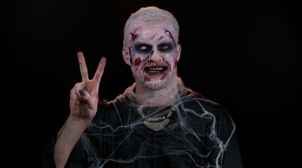 Frightening man with Halloween zombie bloody wounded makeup showing victory sign, hoping for success and win, doing peace gesture, smiling with kind optimistic expression on studio black background