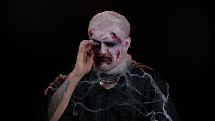 Upset disappointed zombie man with makeup with wounds scars wipes tears and cries from despair,...