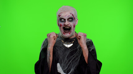 Zombie man with makeup with fake wounds scars celebrate success win scream rejoices doing winner hands gesture say Yes in chroma key studio. Sinister dead guy. Halloween, filming, staging concept