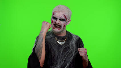 Spooky crazy zombie man alone with makeup with fake wounds scars celebrate success win scream rejoices doing winner hands gesture say Yes in chroma key studio. Sinister dead guy. Halloween holidays
