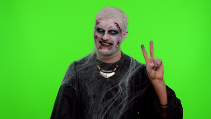 Frightening man with Halloween zombie bloody wounded makeup showing victory sign, hoping for success and win, doing peace gesture, smiling with kind optimistic expression on studio chroma key
