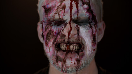 Close-up of sinister man with horrible scary Halloween zombie makeup making faces, looking ominous...