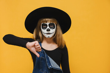 Portrait girl kid with Halloween makeup mask wears black hat giving thumb down gesture looking with negative expression and disapproval, doing bad signal, isolated on yellow wall. Holiday concept