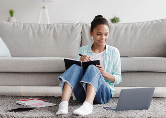 Smiling teen african american girl pupil studies remotely, writes in notebook, sits on floor and looks at laptop