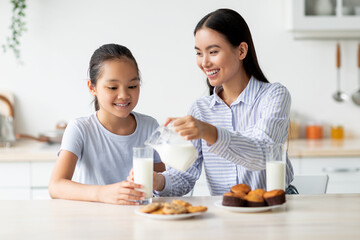 Happy asian mom and daughter eating snacks and drinking milk in kitchen interior, having a bite together