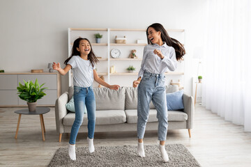Playful korean mother and pretty daughter dancing to music and jumping at home, enjoying favorite song together
