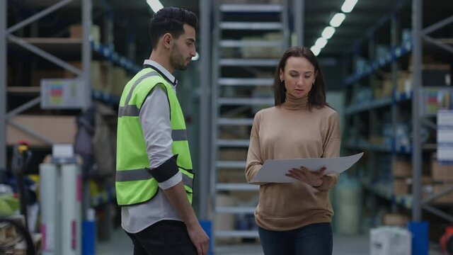 Dissatisfied angry Caucasian woman arguing with Middle Eastern man in warehouse pointing at paperwork. Portrait of female freight agent disappointed with delay problem quarrelling with storage manager