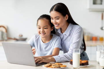 Happy asian mother and kid using laptop together, sitting at kitchen table and looking at computer screen