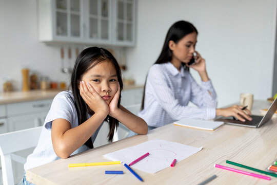 Asian Woman Working On Laptop From Home, Bored Daughter Sitting By Her, Leaning On Her Hands And Looking At Camera