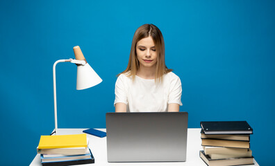 Portrait of a pretty young woman studying while sitting at the table with grey laptop computer, notebook. Smiling business woman working with a laptop isolated on a blue background.