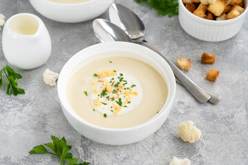 Homemade cauliflower soup puree in a white bowl with cheese, cream, spices and fresh parsley on a gray concrete background. Top view, copy space.