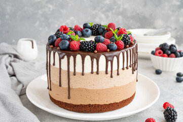 Three chocolate mousse cake with chocolate glaze, blueberries, raspberries and blackberries on top on a white plate on a dark wooden background. Cake with three layers of different taste. Copy space.