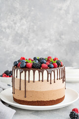 Three chocolate mousse cake with chocolate glaze, blueberries, raspberries and blackberries on top on a white plate on a dark wooden background. Cake with three layers of different taste. Copy space.
