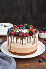 Three chocolate mousse cake with chocolate glaze, blueberries, raspberries and blackberries on top on a white plate on a dark wooden background. Cake with three layers of different taste. Copy space.