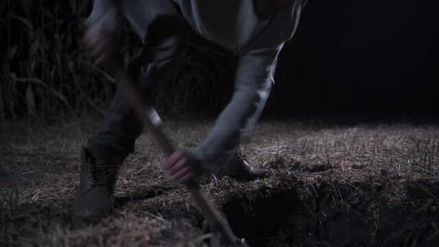 Killer digs grave at night. Young scary man digs hole in field and drinks alcohol. Long shot.