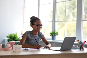 Smiling teen black girl student in glasses studying at home with laptop and watching online lesson