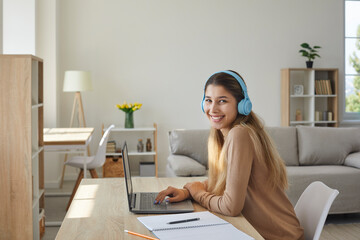 Obraz premium Happy teen girl in headphones at home preparing homework sitting with notebook at laptop. Female student listens to music, does schoolwork at home or searches Internet for information. Side view.