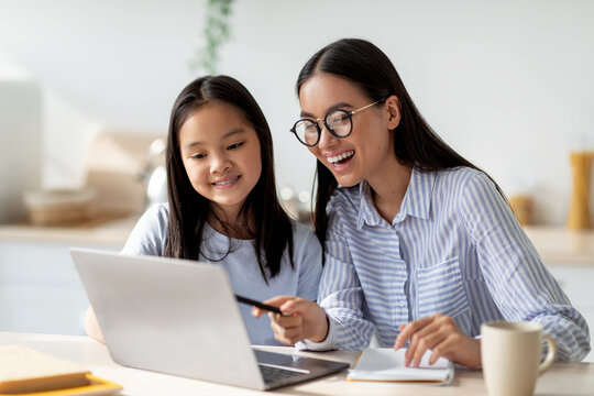 Happy asian mom helping her studying daughter, looking at laptop screen together, sitting in kitchen interior - Powered by Adobe