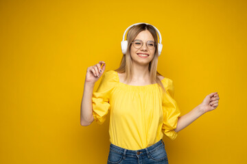 Obraz na płótnie Canvas Beautiful attractive young blond woman wearing yellow t-shirt and glasses in white headphones listening music, dancing and laughing on blue background in studio. Relaxing and enjoying. Lifestyle.