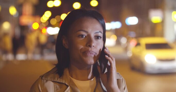 Bokeh shot of mixed-race woman having phone call in evening city with lights in background
