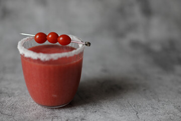 Glass of fresh red tomato juice with salt and metal stick with candy tomatoes on grey background.