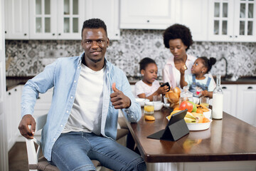 African man smiling, gesturing like and looking at camera while working on digital tablet on kitchen. Pretty woman having breakfast with two daughters on background. Domestic life of beautiful family.