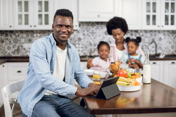 African man smiling and looking at camera while working on digital tablet on bright kitchen. Pretty woman having breakfast with two daughters on background. Domestic life of beautiful family.