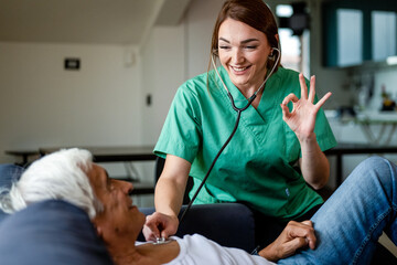 Health visitor using stethoscope to check health of a senior man, young nurse gesturing ok with hand during a home visit, medical health care at home concept