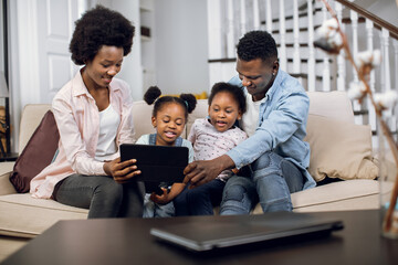 Happy african parents sitting on couch with two pretty daughters and using digital tablet for watching cartoons. Concept of family, leisure time and modern gadgets.